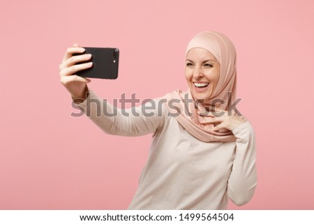 Cheerful young arabian muslim girl in hijab light clothes posing isolated on pink background in studio. People religious Islam lifestyle concept. Mock up copy space. Doing selfie shot on mobile phone