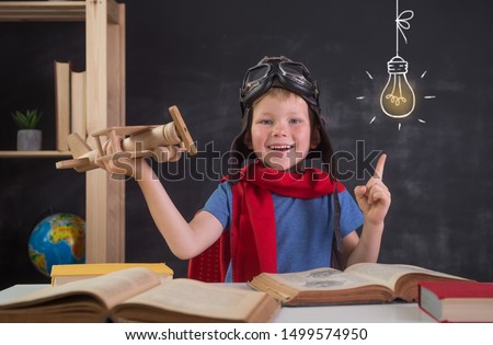 Child sitting the desk and reading a book while playing pilot. Child pilot in helmet and glasses with wooden toy airplane. Innovation and bright idea. Picture of bulb on blackboard