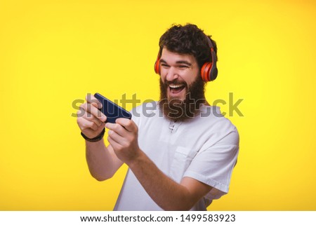Photo of bearded guy playing at mobile phone and using headphones, over yelow backgrdound