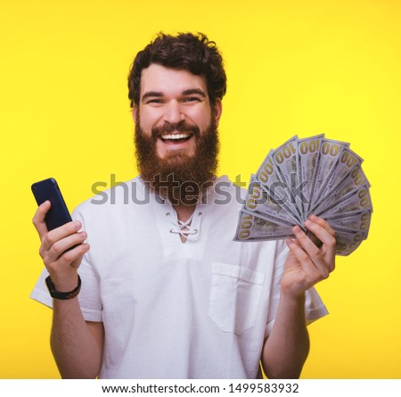 Photo if cheerful bearded man, over yellow background holding mobile phone and a lot of money
