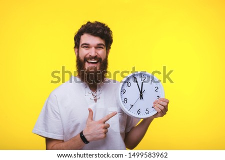 Photo of cheerful bearded man pointing at clock, over isolated background