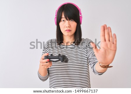 Chinese gamer woman playing video game using headphones over isolated white background with open hand doing stop sign with serious and confident expression, defense gesture
