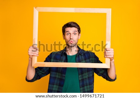 Photo of funny guy holding wooden picture frame looking through it sending air kiss wear casual plaid shirt isolated yellow color background