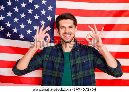 Photo of cool guy standing near usa flag showing okey symbols wear casual plaid shirt isolated on american flag colors background
