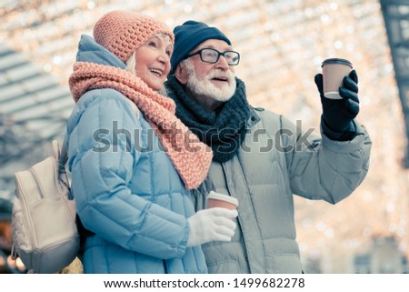 Elderly lady with small backpack standing close to her aged husband and smiling while enjoying coffee to go with him. Christmas illumination on the background