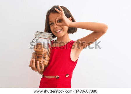 Beautiful child girl holding jar of cookies standing over isolated white background with happy face smiling doing ok sign with hand on eye looking through fingers