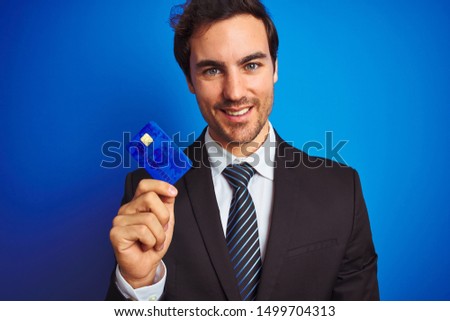 Young handsome businessman holding credit card standing over isolated blue background with a happy face standing and smiling with a confident smile showing teeth