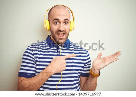 Young man listening to music wearing yellow headphones over isolated background amazed and smiling to the camera while presenting with hand and pointing with finger.