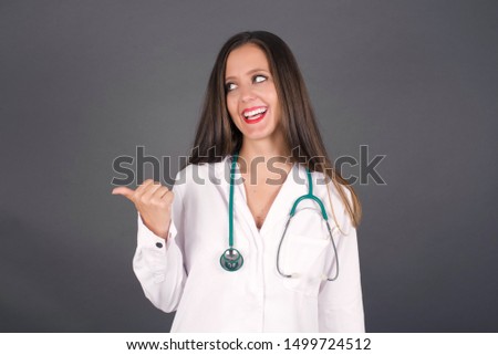 Omg concept. Stupefied caucasian doctor female with surprised expression, opens eyes and mouth widely, points aside with thumb, shows something strange on gray background. Advertisement concept.