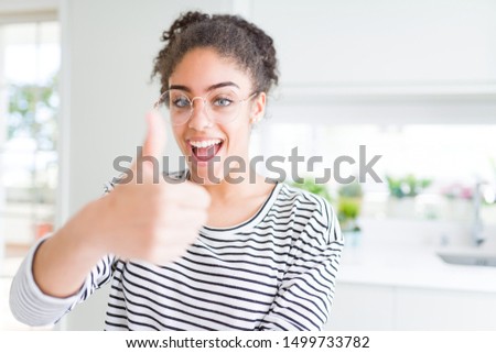 Beautiful young african american woman with afro hair wearing glasses doing happy thumbs up gesture with hand. Approving expression looking at the camera showing success.