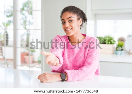 Beautiful african american woman with afro hair wearing casual pink sweater smiling cheerful offering palm hand giving assistance and acceptance.