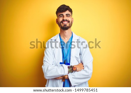 Young doctor man with tattoo wearing id card standing over isolated yellow background happy face smiling with crossed arms looking at the camera. Positive person.