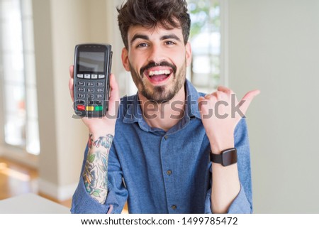Young man holding dataphone point of sale as payment pointing and showing with thumb up to the side with happy face smiling