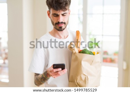 Young handsome man holding a paper bag full of fresh groceries at home, showing and using smartphone while buying products