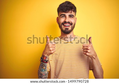 Young man with tattoo wearing striped t-shirt standing over isolated yellow background success sign doing positive gesture with hand, thumbs up smiling and happy. Cheerful expression and winner