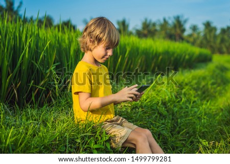 Happy child sitting on the field holding tablet. Boy sitting on the grass on sunny day. Home schooling or playing a tablet