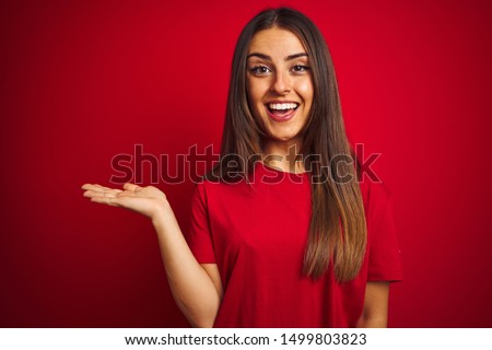 Young beautiful woman wearing t-shirt standing over isolated red background smiling cheerful presenting and pointing with palm of hand looking at the camera.