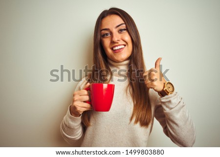 Young beautiful woman drinking red cup of coffee standing over isolated white background happy with big smile doing ok sign, thumb up with fingers, excellent sign