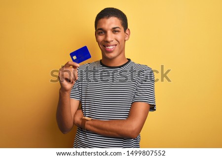Young handsome arab man holding credit card standing over isolated yellow background with a happy face standing and smiling with a confident smile showing teeth