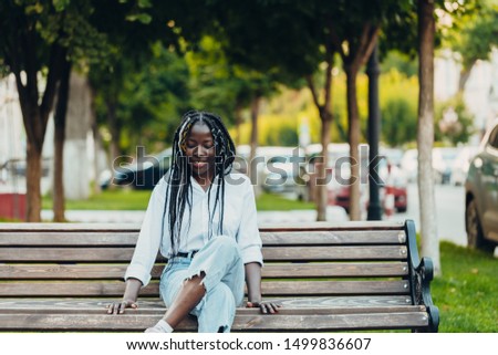 
Close-up portrait of a smiling young African-American girl with pigtails sitting on a bench outside on a sunny day. Outdoor photo.