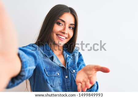 Beautiful woman wearing denim shirt make selfie by camera over isolated white background smiling cheerful offering palm hand giving assistance and acceptance.