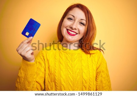 Young customer redhead woman holding credit card over yellow isolated background with a happy face standing and smiling with a confident smile showing teeth