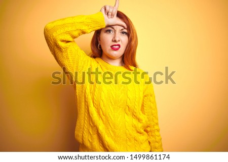 Beautiful redhead woman wearing winter sweater standing over isolated yellow background making fun of people with fingers on forehead doing loser gesture mocking and insulting.