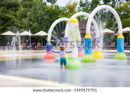 Cute one year blond toddler in swimming suit is playing at water park in sunny summer day, view from back