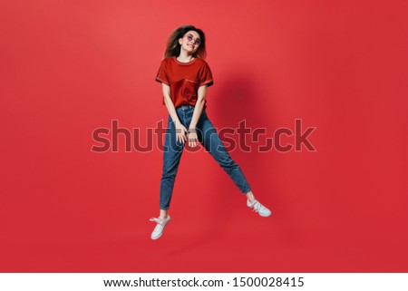 Cute girl in jeans and bright T-shirt jumping on red background