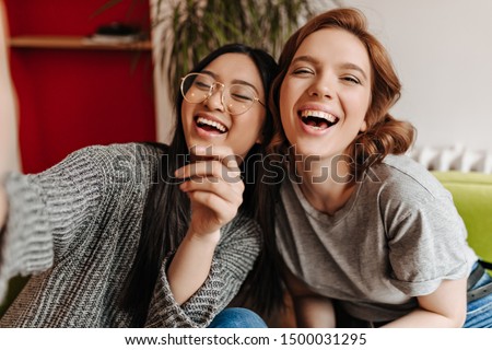 Excited multicultural friends taking selfie and laughing. Indoor shot of two emotional caucasian and chinese girls.