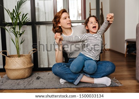 Red-haired woman in striped T-shirt hugs her daughter and plays with her sitting on floor in living room