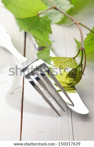 Closeup of silver cutlery with the prongs of a fork and blade of a knife on a white painted wooden background with vine leanes