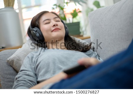 Portrait photo of young Asian woman relaxing and listening to music from smartphone while lying on a sofa.