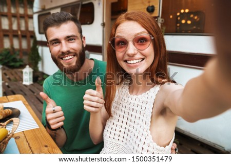 Happy smiling couple having lunch while sitting at the table outdoors, camping at the trailer, taking a selfie, thumbs up