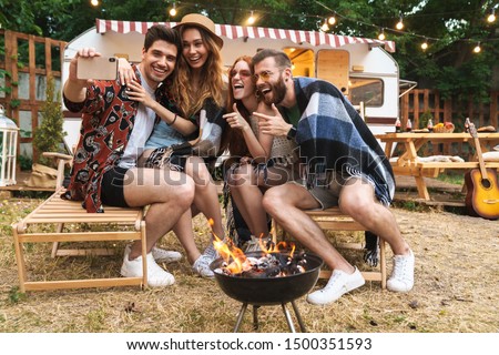 Group of cheerful happy friends sitting at the trailer outdoors, having a picnic with bonfire, taking a selfie