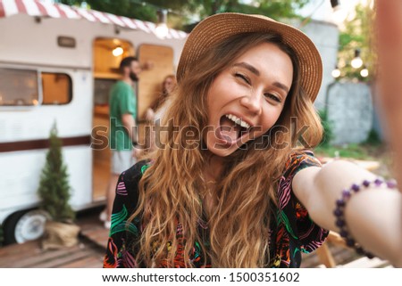 Cheerful young girl taking a selfie while standing at the campsite with a trailer on a background