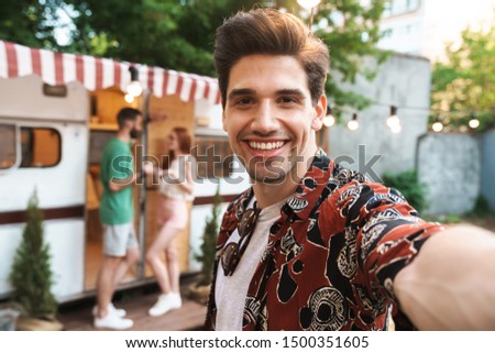 Smiling young man taking a selfie while standing at the campsite with a trailer on a background