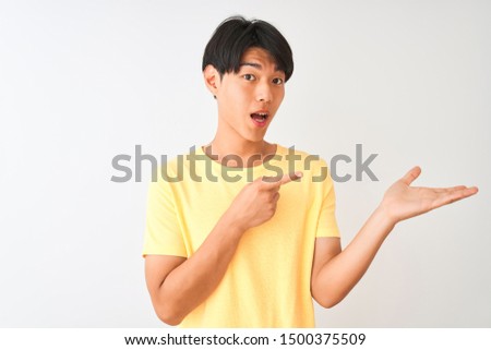 Chinese man wearing yellow casual t-shirt standing over isolated white background amazed and smiling to the camera while presenting with hand and pointing with finger.