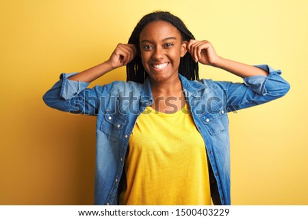 Young african american woman wearing denim shirt standing over isolated yellow background Smiling pulling ears with fingers, funny gesture. Audition problem