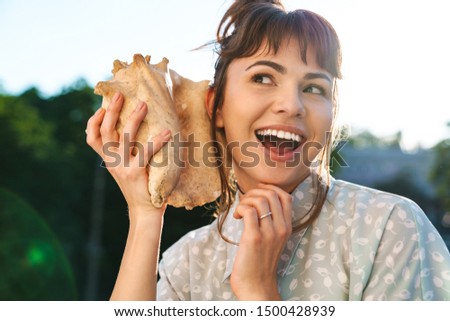 Photo of a cheery laughing young beautiful woman on a balcony holding shell near ear. Royalty-Free Stock Photo #1500428939