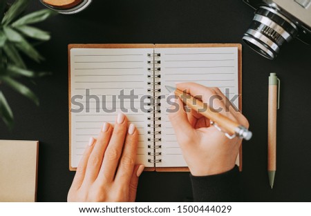 Top view of flat lay woman's hand writes down addresses in an open notebook lying on black table next to camera with pen and notepad. Concept of keeping notes of necessary cases. Copyspace