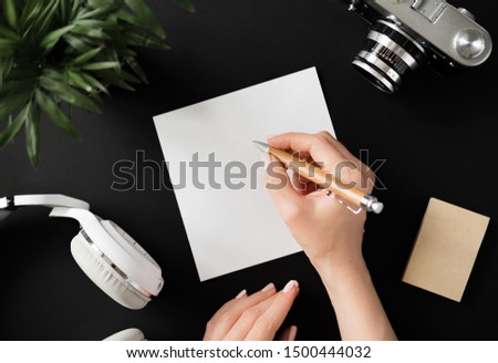Top view of a flat lay female hand writes a note with a pen on a white sheet of paper lying next to the film camera with headphones and recording paper on a black table. Advertising space