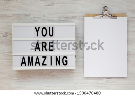 'You are amazing' words on a lightbox, clipboard with blank sheet of paper on a white wooden background, top view. Overhead, from above. Flat lay. Copy space.