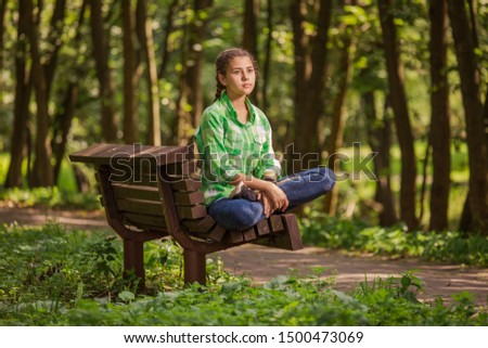 Emotional girl teenager with long hair hairstyle braids in a green shirt sits on a bench in the park.