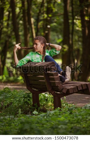 Emotional girl teenager with long hair hairstyle braids in a green shirt sits on a bench in the park.