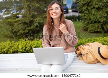 Side view of pleased brunette woman in eyeglasses sitting on bench in park and using laptop computer.Teen girl in white jeans and beige sweater with laptop, backpack and hat.
