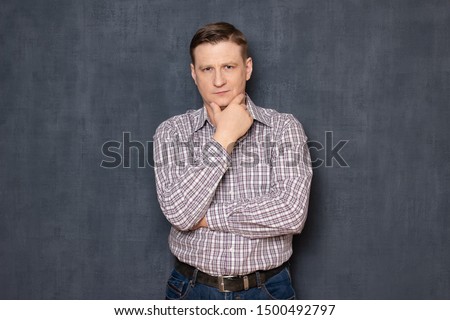 Studio waist-up portrait of thoughtful and confident fair-haired caucasian young man wearing checkered shirt, touching chin with hand, looking with calm expression at camera, over gray background