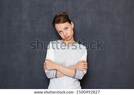 Studio waist-up portrait of tired and bored caucasian fair-haired girl wearing jumper, looking aside and rolling her eyes, pouting lips, crossing hands over chest, standing against gray background