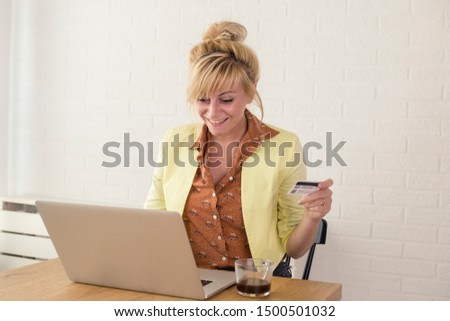 Happy excited woman using credit card and laptop for online shopping. She is sitting at table at home.