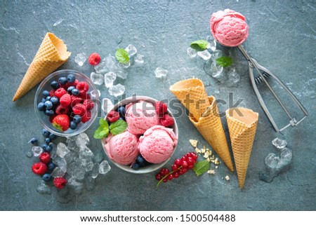 Raspberry ice cream in bowl with berries, ice and scoop on grey stone background